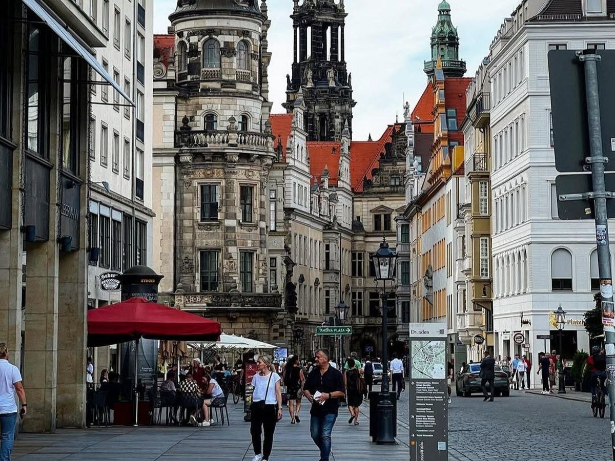 People walking through a historic European old-town street past a café terrace, with baroque towers rising behind them.