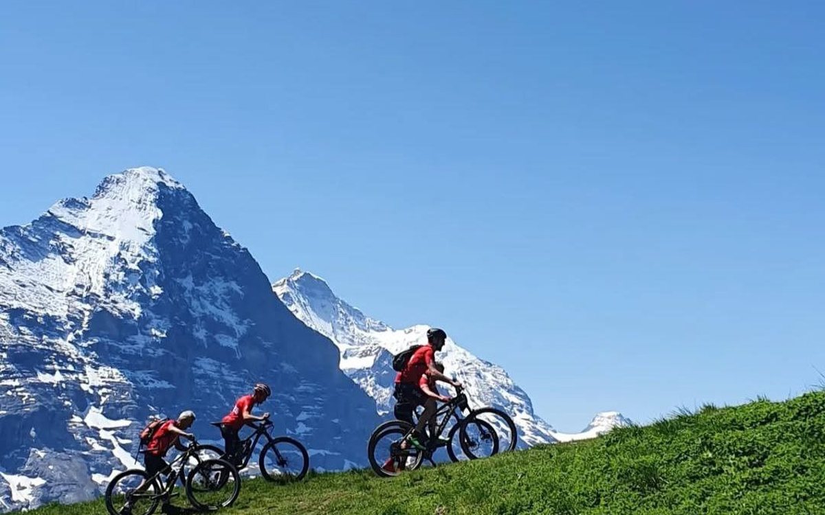 Three cyclists follow a high mountain trail in the Alps, snow-covered peaks rising behind them.